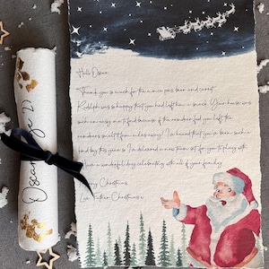 May include: A handwritten letter from Father Christmas to Oscar, featuring a watercolour illustration of Santa Claus and a snowy night sky. The letter thanks Oscar for the mince pies, beer, and carrots and mentions a new train set. The letter is rolled up and tied with a black ribbon.