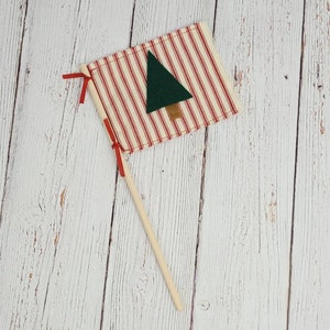 May include: A red and white striped fabric flag with a green felt Christmas tree applique. The flag is attached to a wooden stick.