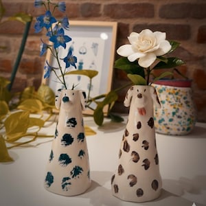 Può includere: Due vasi in ceramica a forma di cane, uno con accenti floreali blu e l'altro con macchie marroni, ciascuno con fiori. I vasi sono bianchi con segni neri e marroni, su uno sfondo di muro di mattoni.
