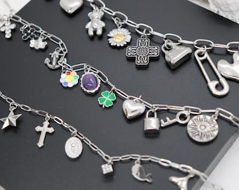 Könnte beinhalten: Drei silberfarbene Metall-Charm-Armbänder mit einer Vielzahl von hängenden Anhängern, darunter ein Teddybär, ein Gänseblümchen, ein Kreuz, eine Sicherheitsnadel, ein Herz, ein Dolch, eine Erdbeere, ein Stern, ein violetter ovaler Stein, ein Kleeblatt und eine Sonne.