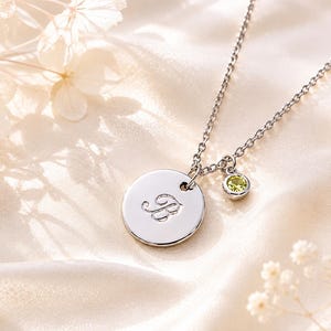 May include: A silver-toned metal necklace featuring a circular pendant engraved with the cursive letter B and a smaller, round, light green gemstone charm attached to the delicate chain link, creating a simple and elegant jewellery piece.