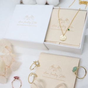 以下が含まれることがあります： 「Anna Jewellery Studio」と刻まれた金色のロゴがついた白い箱と、緑色の石のペンダントがついた金色のネックレス。ネックレスは、ベージュの裏地が付いた白い箱に入っています。ベージュの表面には、金色の指輪がいくつか置かれています。