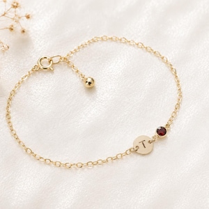 May include: A delicate gold chain bracelet featuring a small circular charm engraved with the letter T and a single round dark red gemstone. The bracelet includes a spring ring clasp and a short extender chain finished with a small gold bead.