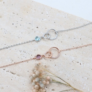 Könnte beinhalten: Zwei zarte Armbänder mit ineinandergreifenden Kreisen, die mit kleinen Edelsteinen verziert sind. Ein Armband ist silber mit einem hellblauen Edelstein, das andere ist roségold mit einem rosafarbenen Edelstein.