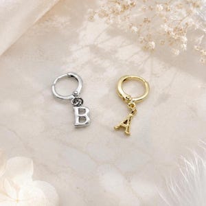 May include: Two small hoop earrings, one in a silver tone featuring a dangling letter B charm and one in a gold tone featuring a dangling letter A charm. Each earring has a hinged closure mechanism and a polished metallic finish on the letters.