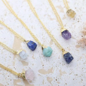 May include: A collection of necklaces featuring raw, uncut gemstones in various colors including blue, purple, green, and pink, each suspended from a thin gold-tone metal chain with a gold-tone cap securing the stone to the chain.