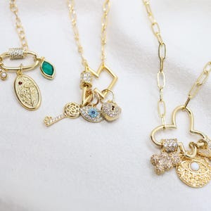 Pode incluir: Três colares dourados com vários pingentes. Um apresenta uma lua crescente, um oval com um design floral e uma gema verde. Outro tem uma chave, um olho e um cadeado em forma de coração. O terceiro tem um pingente em forma de trevo.