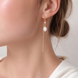 May include: A gold threader earring featuring a small white pearl and a delicate chain that hangs down. The earring is constructed with a thin gold wire that passes through the earlobe, suspending a single teardrop-shaped pearl accent.
