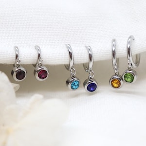 May include: Three pairs of silver-toned hoop earrings, each featuring a small circular charm set with a faceted gemstone. The stones are coloured deep red, light blue, dark blue, orange, and bright green, arranged in a row on a white fabric.