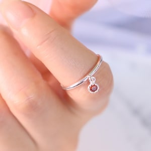 Op de afbeelding: Een dunne zilveren ring met een kleine, ronde bedel die aan de ringband hangt. De bedel is gezet met een enkele, ronde, dieprode edelsteen, omringd door een delicate halo van piepkleine, heldere, fonkelende steentjes die het licht vangen.