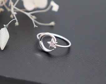 May include: A silver-toned ring featuring a crescent moon motif and a small five-pointed star. The star is positioned within the curve of the moon and contains a single, small, clear embedded stone at its center. The band is thin and circular.