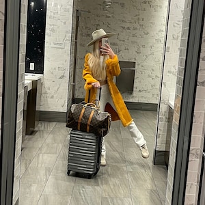 Op de afbeelding: Een vrouw in een gele trui en een witte hoed staat in een badkamer met een koffer. Ze houdt haar telefoon vast en maakt een selfie.