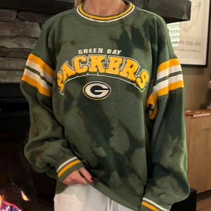 May include: Green Bay Packers sweatshirt in dark green with yellow and white stripes on the sleeves. The team name "PACKERS" is in yellow block letters across the chest, with the team logo below. The sweatshirt has a crew neck and ribbed cuffs.