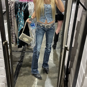 Puede incluir: Un espejo de cuerpo entero refleja a una persona con un top de mezclilla, jeans azules y un sombrero de vaquero negro. La persona sostiene una botella de agua y un pequeño bolso estampado. Se ven botas de vaquero en el reflejo.