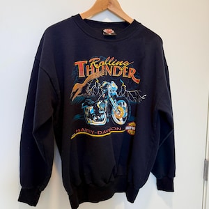 Puede incluir: Sudadera negra con un gráfico de una motocicleta Harley-Davidson y el texto "Rolling Thunder" en naranja y amarillo. El texto "Harley-Davidson" está impreso debajo de la motocicleta.