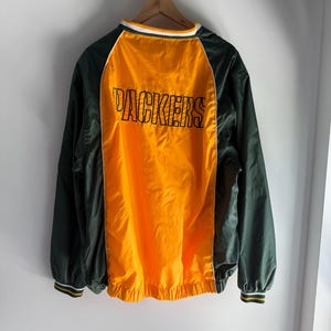 Könnte beinhalten: Eine grün-goldene Jacke mit dem Wort "PACKERS" auf dem Rücken. Die Jacke hat eine gelbe Mittelbahn und grüne Ärmel. Kragen und Manschetten haben weiße und gelbe Streifen.