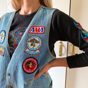 May include: A blue denim vest with multiple patches, including an AMA Motorcycle Rally & Race patch, a Black Hills Motorcycle Rally & Race patch, and a Sturgis SD Centennial patch. The vest is worn over a black long-sleeve shirt with a flame design.