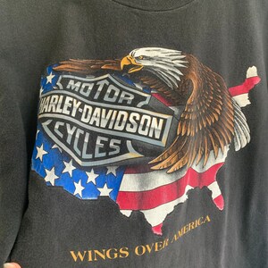 May include: Black t-shirt with a graphic of a bald eagle flying over a map of the United States. The eagle is holding a Harley-Davidson logo in its talons. The text "MOTOR HARLEY-DAVIDSON CYCLES" is on the logo. The text "WINGS OVER AMERICA" is below the graphic.