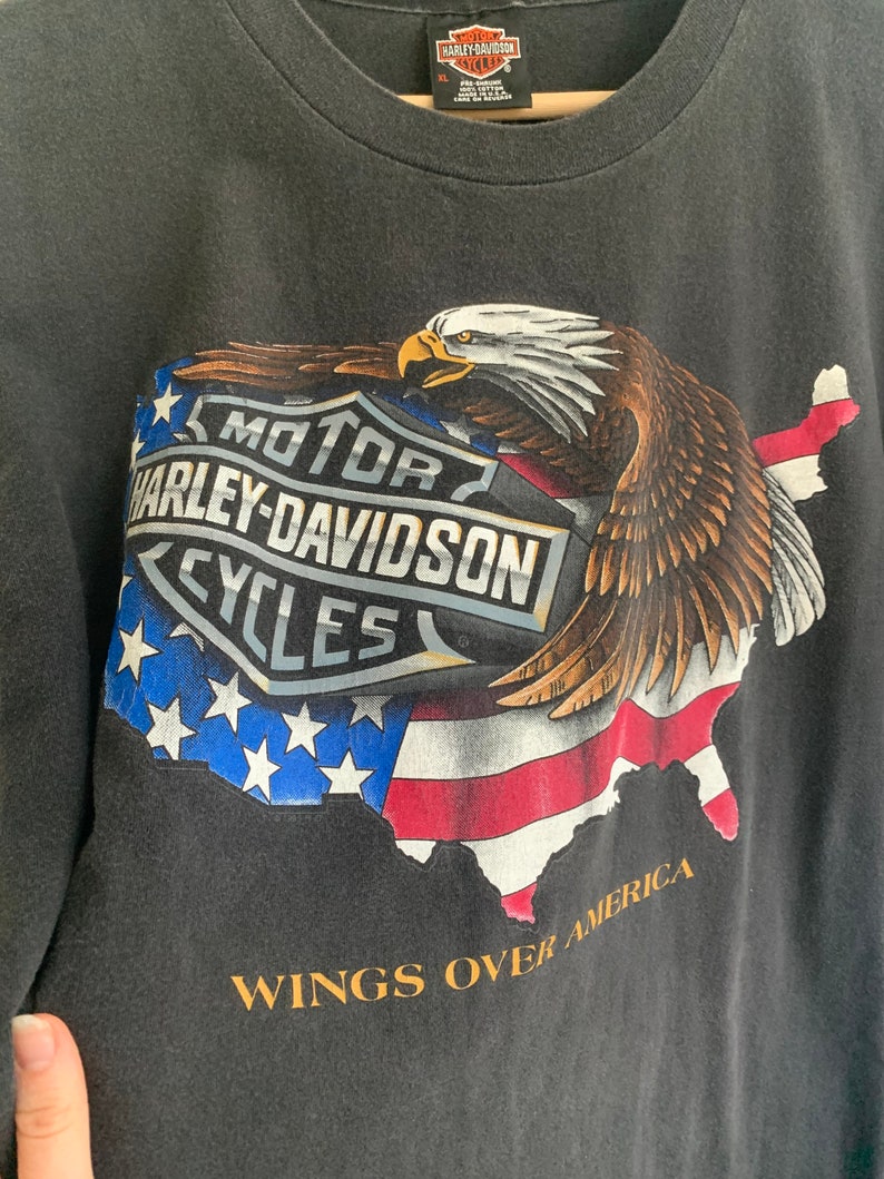 May include: Black t-shirt with a graphic of a bald eagle flying over a map of the United States. The eagle is holding a Harley-Davidson logo in its talons. The text "MOTOR HARLEY-DAVIDSON CYCLES" is on the logo. The text "WINGS OVER AMERICA" is below the graphic.
