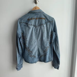May include: Light blue denim jacket with long sleeves, featuring the Harley-Davidson logo embroidered across the upper back. The jacket has decorative stitching and stud accents. The jacket is hanging on a wooden hanger.