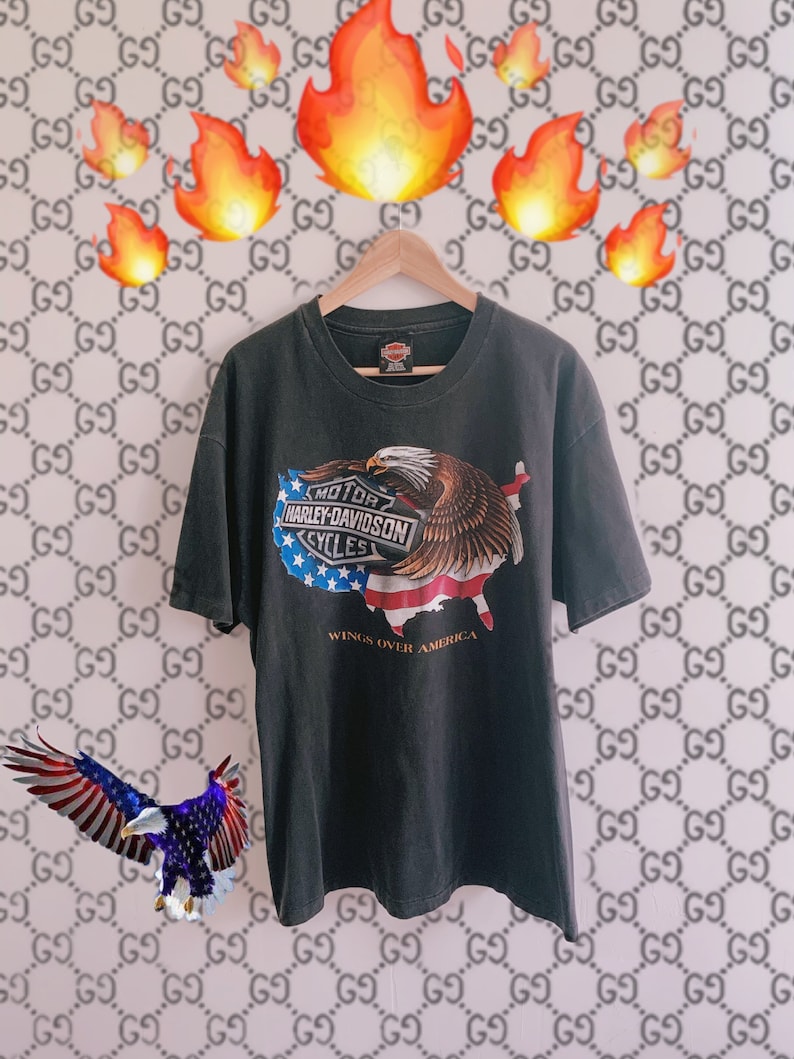 May include: A black t-shirt with a Harley Davidson logo and an American eagle graphic. The eagle is flying over a map of the United States. The text 'Wings Over America' is printed below the graphic. The t-shirt is hanging on a wooden hanger against a white background with a repeating pattern of the letter G.