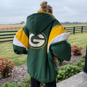 May include: A dark green hooded jacket featuring a large yellow and white letter G logo on the back. The jacket has quilted yellow panels on the shoulders and white accents on the sleeves, with a textured fabric finish and a drawstring hood.