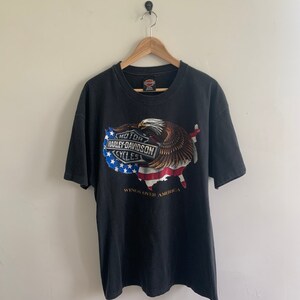 May include: Black t-shirt with a Harley-Davidson logo and an American eagle graphic. The eagle is holding a US flag in its talons. The text "Wings Over America" is printed below the graphic.