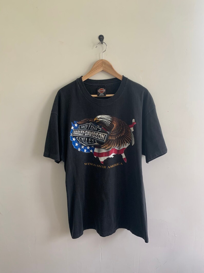 May include: Black t-shirt with a Harley-Davidson logo and an American eagle graphic. The eagle is holding a US flag in its talons. The text "Wings Over America" is printed below the graphic.