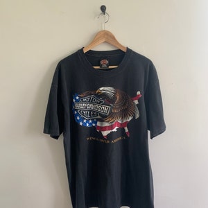 May include: Black t-shirt with a graphic of an eagle flying over a stylized American flag. The graphic includes the Harley-Davidson logo and the text "Wings Over America".