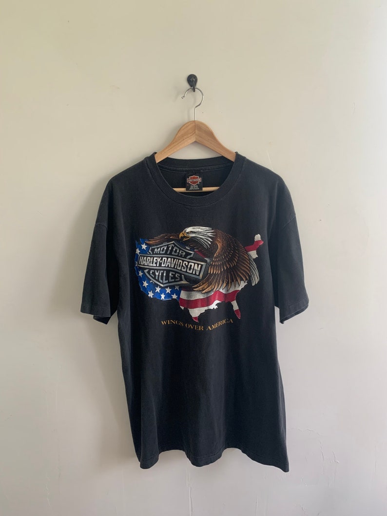 May include: Black t-shirt with a graphic of an eagle flying over a stylized American flag. The graphic includes the Harley-Davidson logo and the text "Wings Over America".
