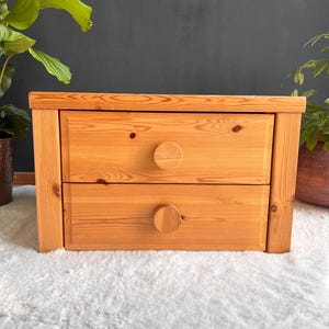 Może przedstawiać: Dwuszeregowa drewniana szafka nocna z naturalnym wykończeniem. Szafka nocna ma okrągłe drewniane gałki i prosty, prostokątny kształt. Widoczne jest usłojenie drewna, a mebel stoi na ciemnym tle.