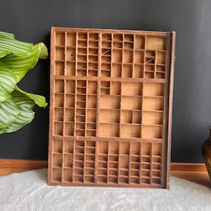 May include: A vintage wooden printer's tray with many small compartments. The tray is made of light brown wood and is rectangular. It is designed to organise small items or display collectibles. The background is a dark wall and a white fluffy rug.