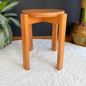 May include: A round wooden stool featuring a light brown grain finish, constructed with three thick, vertical legs that support a circular seat. The stool has a simple, structural design with visible wood grain patterns throughout the entire piece.
