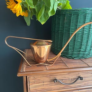 Puede incluir: Un calentador de té de color cobre con un asa curva y una tapa pequeña y redonda. El calentador se encuentra sobre un soporte de metal dorado. Una cesta de mimbre verde y girasoles están en el fondo. El calentador de té está sobre una cómoda de bambú.