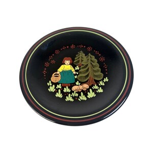 May include: Black decorative plate featuring a painted scene of a girl with a basket, trees, and mushrooms. The plate has a brown and yellow striped border. The girl wears a yellow top and green skirt.