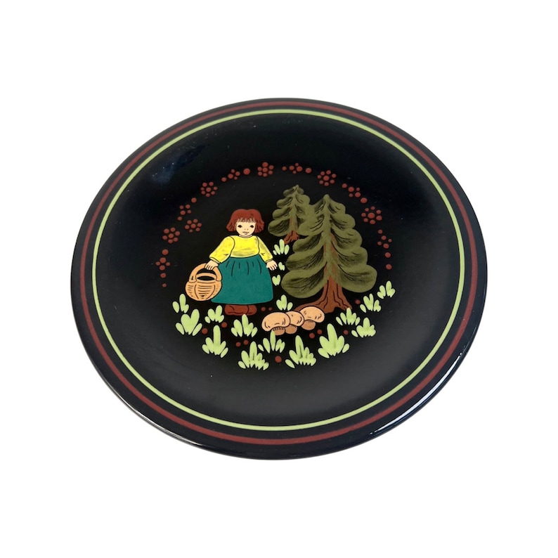 May include: Black decorative plate featuring a painted scene of a girl with a basket, trees, and mushrooms. The plate has a brown and yellow striped border. The girl wears a yellow top and green skirt.