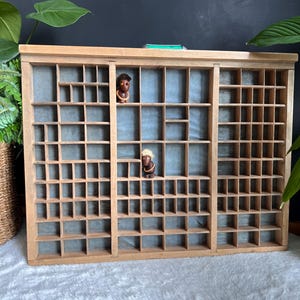 May include: A wooden printer's tray with many small compartments, suitable for displaying collectibles. Two small figurines are placed inside the tray. The tray is made of light-coloured wood and is rectangular. The background is a dark colour.
