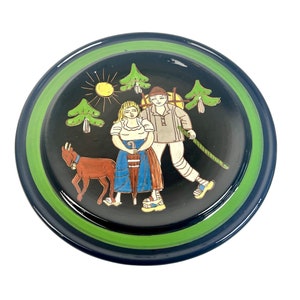 May include: A round ceramic plate with a black background and green trim. The plate features a painted scene of a couple in traditional clothing walking with a goat in a forest setting. The scene includes trees, a sun, and a blue umbrella.