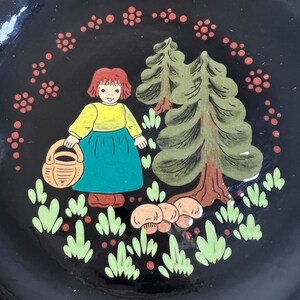May include: A black decorative plate with a hand-painted illustration. The image features a girl in a yellow top and blue skirt holding a basket, with trees and mushrooms. The plate is surrounded by a ring of red floral accents.