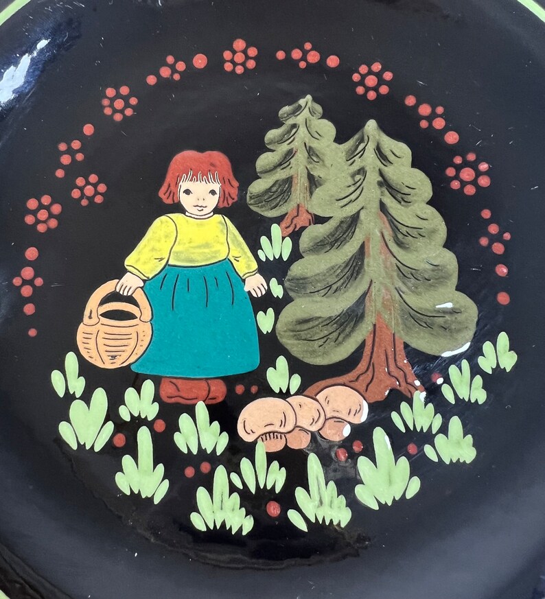 May include: A black decorative plate with a hand-painted illustration. The image features a girl in a yellow top and blue skirt holding a basket, with trees and mushrooms. The plate is surrounded by a ring of red floral accents.