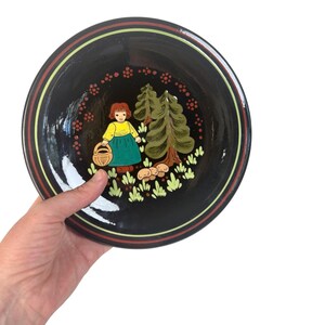 May include: A black decorative plate featuring a hand-painted illustration of a girl with a basket, trees, and mushrooms. The plate has a glossy finish and a striped border in brown, yellow, and black. The artwork is in a folk art style.