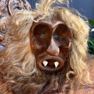 Könnte beinhalten: Eine geschnitzte Holzmaske mit detailliertem Gesicht, großen Augenhöhlen und zwei weißen Zähnen. Die Maske hat eine hellbraune Farbe und ist von langen, hellbraunen Haaren umgeben. Die Maske ist wahrscheinlich ein dekoratives oder zeremonielles Objekt.