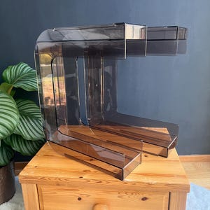 Puede incluir: Un organizador de escritorio acrílico, claro y angular, con un diseño escalonado. El organizador es de color gris ahumado y se encuentra sobre una mesita de noche de madera. El diseño presenta una serie de compartimentos en forma de L.