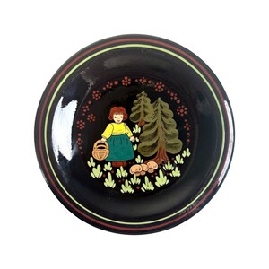 May include: A round, black decorative plate featuring a painted scene of a girl in a yellow top and green skirt holding a basket, near trees and mushrooms. The plate has a brown and yellow striped border.