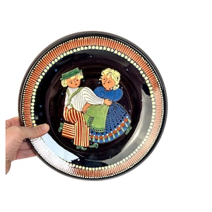 May include: A round ceramic plate with a black background and a colorful illustration of a couple dancing. The plate has a brown and orange border with white dots.