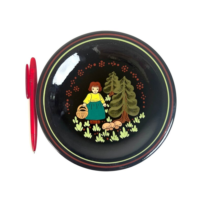 May include: A black decorative plate featuring a painted scene of a girl in a yellow top and green skirt holding a basket, near trees and mushrooms. The plate has a red, yellow, and brown border. A red pen is next to the plate.