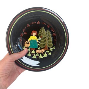 May include: Black ceramic plate featuring a painted scene of a girl with a basket, trees, and mushrooms. The plate has a red, yellow, and black border. The girl wears a yellow top and green skirt. The trees are green and brown.