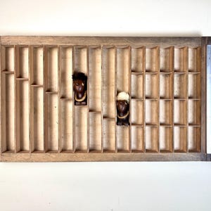 Puede incluir: Un bloque de impresión de madera con dos pequeñas figuras de madera en los compartimentos. Las figuras son marrones y negras con pelo blanco.