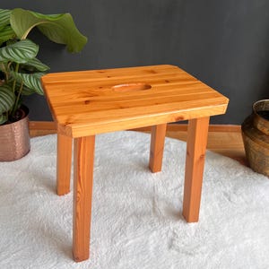 Op de afbeelding: Een kleine, vierkante houten kruk met vier poten. De kruk is gemaakt van lichtgekleurd hout en heeft een rechthoekige uitsparing in het midden van de zitting. De kruk staat op een wit, pluizig tapijt.