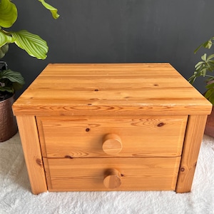 May include: A small, two-drawer wooden chest with a natural finish. The chest has round wooden knobs and is made of light-colored wood. The drawers are rectangular and the chest is sitting on a white, fluffy surface.